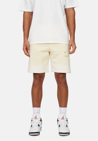 Pantaloni corto in cotone beige con una vestibilità rilassata, dotati di un logo stampato in verde sulla gamba sinistra. Indossati con sneakers bianche e calzini bianchi.