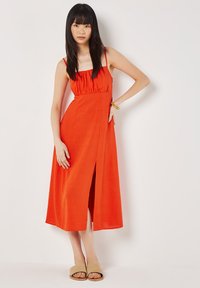 Maxikleid aus leuchtend orangefarbenem Stoff mit verstellbaren Spaghettiträgern, tailliertem Schnitt und einem seitlichen Schlitz. Kombiniert mit hellen flachen Sandalen.