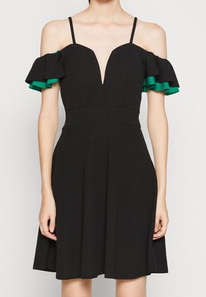 Zwarte off-shoulder jurk met dunne bandjes, diepe V-halslijn en gelaagde groene ruches aan de mouwen bij een staande persoon.