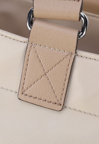 Dettaglio di una borsa in tessuto beige che mostra una tracolla in pelle beige chiaro, liscia, con un accento cucito a forma di X e un attacco in metallo argentato.