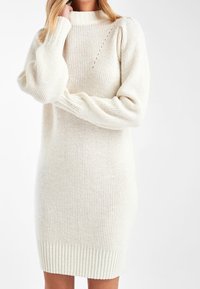 Femme portant une robe pull en maille de couleur crème avec des manches longues bouffantes et des poignets côtelés, debout devant un fond blanc.