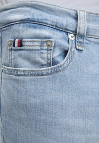 Κοντινή λήψη τσέπης τζιν ανοιχτού μπλε με μικρή υφασμάτινη ετικέτα με κόκκινες, λευκές και μπλε ρίγες και μεταλλικά καρφιά.
