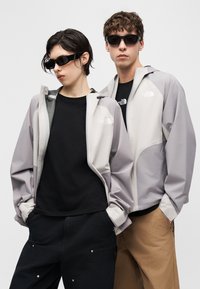 Δύο άτομα φορούν ανοιχτό γκρι μπουφάν North Face πάνω από μαύρα πουκάμισα, μαύρα γυαλιά ηλίου, με τα χέρια στις τσέπες τους μπροστά σε ένα απλό ανοιχτό φόντο.