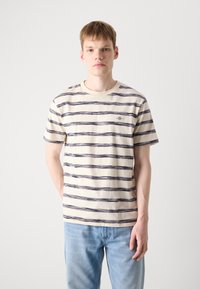 Tricou din bumbac cu dungi, având o culoare de bază crem și dungi orizontale de culoare albastru închis. Guler rotund și mâneci scurte. Detaliu subtil al logo-ului.