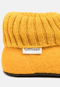 Gelbe Hausschuhe mit einem gerippten gestrickten Oberteil und einem weichen Filzkörper, einer schwarzen Sohle und einem weißen Etikett mit der Aufschrift "Gottstein."