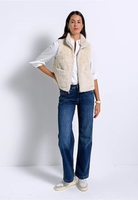 Gilet in pelliccia sintetica color crema, con chiusura frontale a zip, due tasche frontali e colletto, indossato sopra una camicia bianca e jeans blu.