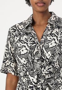 Abito camicia stampato floreale in bianco e nero con colletto, maniche corte e dettaglio in vita legato. Tessuto liscio con stampa testurizzata.