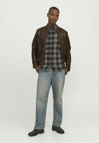 Braune Lederbomberjacke mit gesteppten Akzenten, schwarz-weiße Karohemd, hellblaue weite Jeans und schwarze Schuhe.