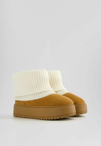 Bottes cheville en daim marron avec un épais bord côtelé en tricot crème et une semelle plateforme texturée, présentant un design à bout arrondi.