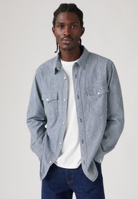 Ριγέ μπλε και λευκό πουκάμισο με κουμπιά και δύο τσέπες στο στήθος. Φορεμένο πάνω από λευκό t-shirt, σε συνδυασμό με σκούρο μπλε τζιν. Λείο υφής.