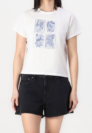 Kobieta ubrana w białą krótką bluzkę z niebieskim nadrukiem owoców i czarne krótkie spodenki z wysokim stanem, stojąca na jednolitym tle.