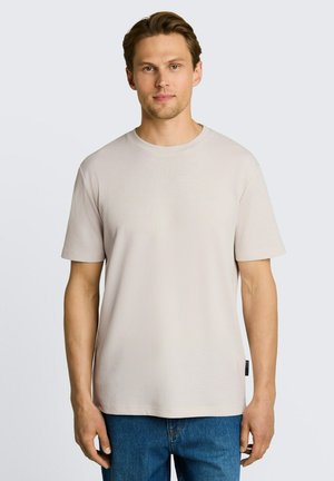 Junger Mann mit hellbraunem Haar, der ein beiges Kurzarmhemd und blaue Jeans trägt, steht vor einem schlichten hellen Hintergrund.