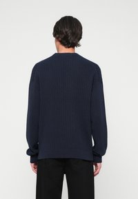Pulover bleumarin cu model cu nervuri, cu decolteu rotund și mâneci raglan, având o croială lejeră și o suprafață texturată. Pantaloni negri vizibili în partea de jos.