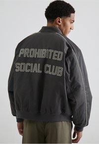 Jachetă bombă gri cu textul "PROHIBITED SOCIAL CLUB" brodat pe spate în alb. Prezintă un guler cu dungi și manșete elastice.