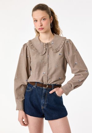 Ung kvinde med krøllet hår i en hestehale iført en beige stribet bluse med flæsekant og blå denimshorts med et brunt bælte.