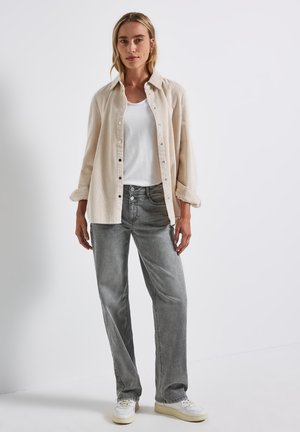 Beige Corduroy-Hemd mit Knöpfen über einem weißen T-Shirt, kombiniert mit grauen Jeans im Straight-Leg-Schnitt und weißen Sneakern, vor einem schlichten Hintergrund.
