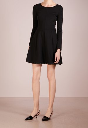 Robe noire à manches longues avec un corsage ajusté et une jupe évasée. Présente des clous décoratifs le long de l'ourlet. Portée avec des talons slingback noirs.
