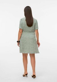 Robe verte à manches courtes, motif floral, taille cintrée et jupe évasée. Portée avec des sandales à talons bruns.