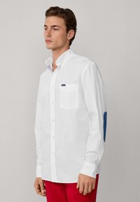 Chemise blanche à boutons avec une poche poitrine, coudières contrastantes en bleu et un col rayé à l'intérieur. Fabriquée en tissu lisse, au design ajusté.