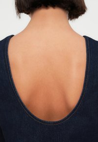 Τζιν φόρεμα με βαθύ, στρογγυλεμένο άνοιγμα στην πλάτη, ραφές ανοιχτού καφέ χρώματος, αντίθετη υφή υφάσματος που αναδεικνύει τον τόνο του δέρματος.