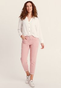 Chemise blanche à manches longues, associée à un pantalon court rose clair avec taille à cordon et poches latérales. Baskets blanches décontractées.
