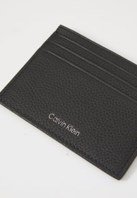 Card holder din piele neagră texturată cu trei sloturi pentru carduri și logo-ul argintiu Calvin Klein pe un fundal alb.