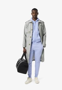 Grau karierten Trenchcoat mit goldenen Knöpfen, über einem hellblauen Hoodie und passenden Jogginghosen getragen. Eine schwarze Reisetasche und cremefarbene Sneakers runden das Outfit ab.