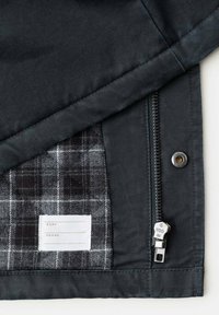 Coin de veste noire montrant un tissu intérieur en plaid gris, une fermeture éclair en métal, un bouton pression et une étiquette blanche avec des champs pour le nom et le numéro de téléphone.