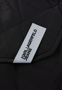 Sort quiltet stof med en glat tekstur; har et lysegråt mærke med "KARL LAGERFELD JEANS" præget i sort.