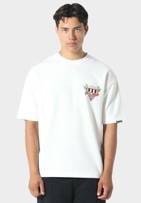 Hvid bomulds T-shirt med en løs pasform, der har et farverigt grafisk design med palmer og geometriske mønstre på brystet.