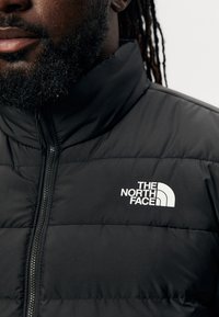 Zwarte pufferjack met een hoge kraag en een gevoerde structuur. Heeft een wit logo op de linkerkant van de borst en een ritsafsluiting.