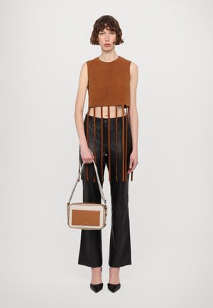 Top senza maniche in suede marrone con frange, abbinato a pantaloni neri a campana. Tiene una borsa rettangolare con sezioni a motivi e di un colore uniforme.