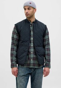 Gilet nero trapuntato con colletto alto, indossato sopra una camicia a quadri verde e bordeaux. Il tessuto è liscio e presenta tasche laterali. Incluso un berretto lavorato a maglia grigio.