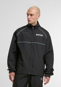 Veste coupe-vent noire avec un col haut, fermeture éclair sur le devant et des accents de couture bleu clair. Le tissu a une finition légèrement texturée.