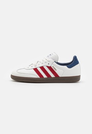Zapatilla Adidas Samba blanca con rayas rojas, lengüeta trasera azul marino, suela marrón y cordones blancos, vista desde un lado sobre un fondo liso.