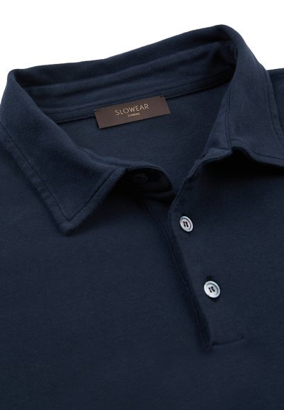 Polo bleu marine à col avec deux boutons blancs, comportant une étiquette noire portant la mention "SLOWEAR ZAMONE" à l'intérieur du col.