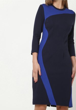 Femme portant une robe noire moulante longueur genou avec des panneaux courbes bleus et des manches trois-quarts, debout devant un fond clair uni.