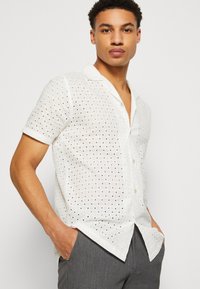 White short-sleeve shirt with a loose fit, featuring eyelet pattern and two front pockets. Soft, lightweight fabric with a relaxed design.