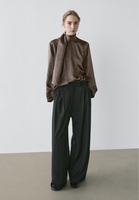 Blouse en satin marron avec un nœud au col, associée à un pantalon large noir taille haute. Texture lisse et design minimaliste.