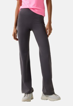 Kobieta ubrana w ciemnoszare spodnie o szerokich nogawkach, różowy top i jasnoszare masywne buty sportowe, stojąca na jasnym, jednolitym tle.