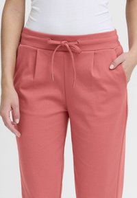Rosa elastiska byxor med dragsko i midjan, med veck framtill, tillverkade av mjukt tyg, och sidofickor för bekvämlighet.
