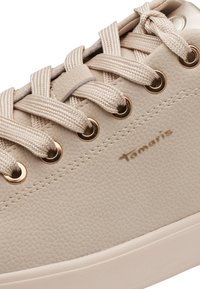 Gros plan d'une basket beige en cuir texturé, lacets en tissu passés dans des œillets métalliques, et logo Tamari embossé sur le côté.