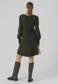 Robe-pull en maille vert foncé, dotée de manches longues bouffantes et d'un ourlet évasé. Associée à des bottes en cuir marron montant jusqu'aux genoux et un petit sac à main.