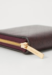 Calvin Klein wallet in burgundy textured material, featuring a gold zipper and logo pull, with a contrasting fabric accent.