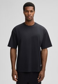 Mann in schlichtem schwarzen, übergroßen T-Shirt mit kurzen Ärmeln und schwarzen Hosen, steht vor einem hellgrauen Hintergrund.