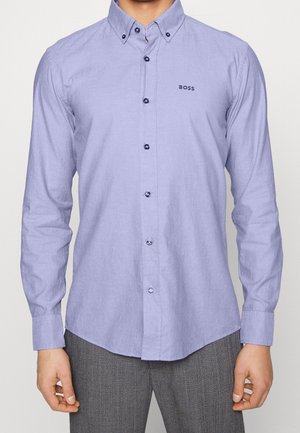 Mężczyzna ubrany w jasnofioletową koszulę zapinaną na guziki z logo "BOSS" na lewej piersi oraz szare spodnie o fakturze.