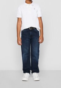 Ein Kind trägt ein weißes Poloshirt, dunkelblaue Jeans mit einem schwarzen Gürtel und weiße Turnschuhe. Es steht vor einem schlichten hellen Hintergrund.