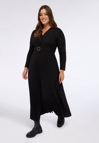 Rochie neagră cu mâneci lungi, cu design înfășurat, featuring un tiv texturat, fustă pliată și un accent din curea circulare. Asortată cu cizme negre până la glezne.
