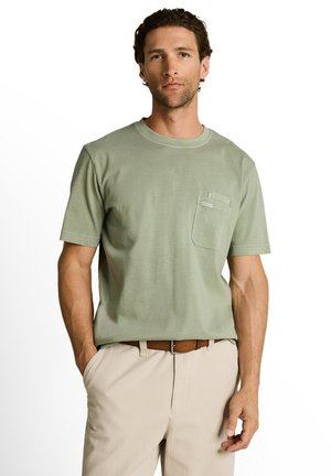 Man draagt lichtgroen T-shirt met korte mouwen en borstzak en beige broek, staat met één hand in de zak tegen een witte achtergrond.