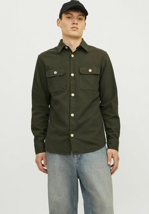Camicia button-up verde scuro con due tasche sul petto e bottoni di colore chiaro, abbinata a jeans azzurri. Tessuto dalla texture liscia.